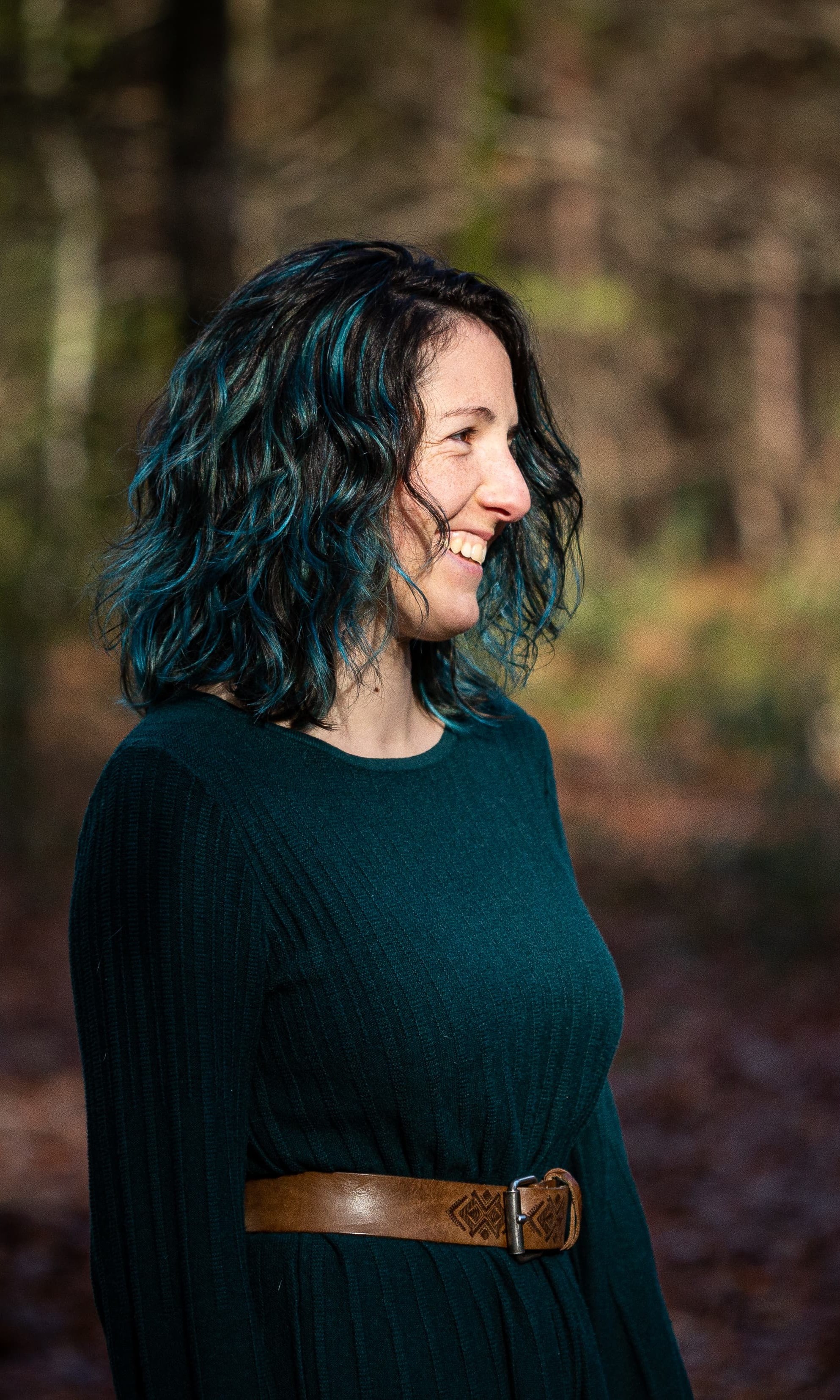 Photographie de moi, debout, de profil, en train de faire un grand sourire sincère. Je porte une robe bleue émeraude et une ceinture brune.